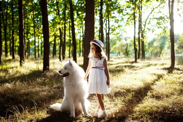 Niña bonita en vestido y sombrero caminando, jugando con un perro blanco en el parque al atardecer.