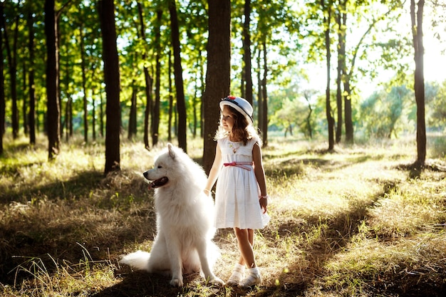 Niña bonita en vestido y sombrero caminando, jugando con un perro blanco en el parque al atardecer.