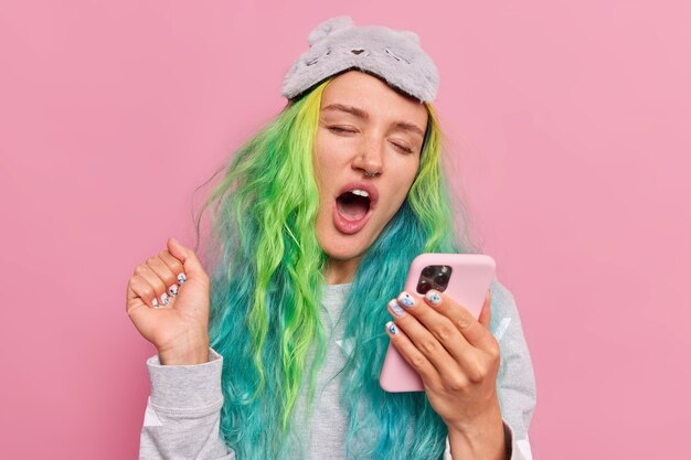 La niña bosteza y navega por internet a través de un celular después de despertar mantiene la mano levantada usa ropa de dormir poses de máscara de dormir en rosa