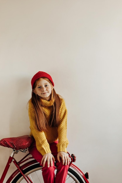 Niña con cabello largo y pecas en suéter amarillo gorra roja y pantalones sentada en bicicleta sobre fondo blanco aislado