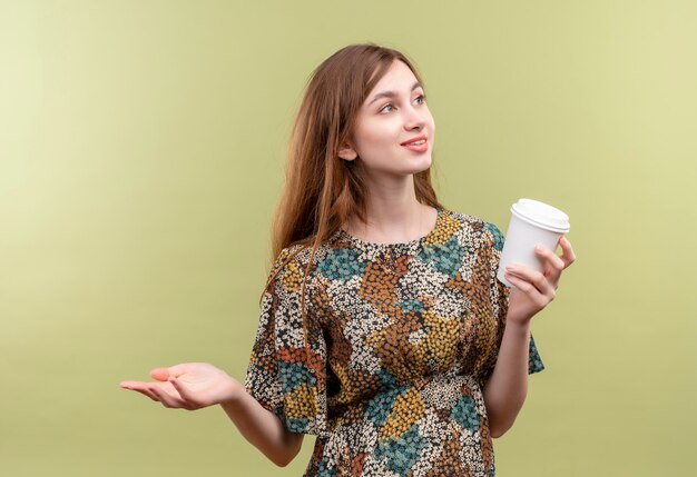 Niña con cabello largo vistiendo coloridos vestidos sosteniendo la taza de café sonriendo mirando a un lado extendiendo el brazo hacia el lado