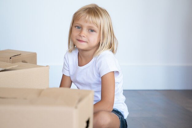La niña de cabello rubio más linda positiva sentada en el piso cerca de cajas de dibujos animados en el nuevo apartamento y mirando adentro