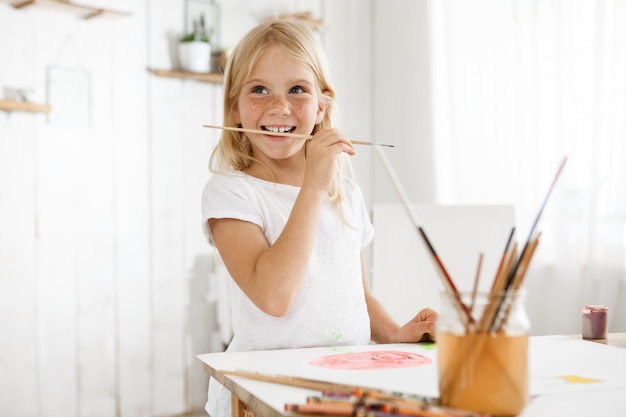 Niña con cabello rubio y pecas disfrutando de arte con camiseta blanca. Niña capturada por un impulso creativo que muerde el cepillo. Niños, arte y emociones positivas.