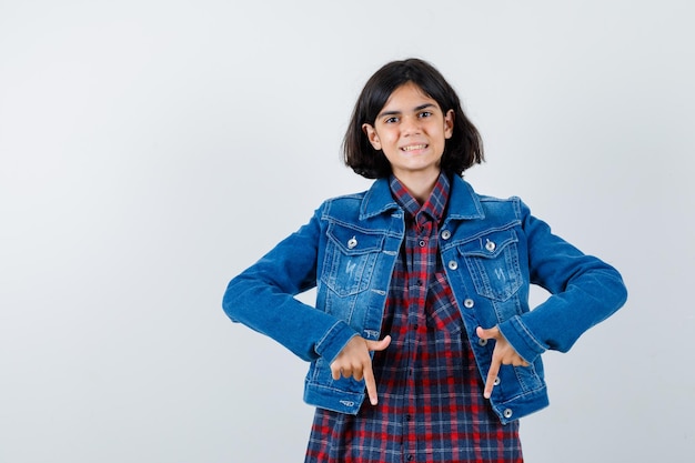 Niña en camisa, chaqueta apuntando hacia abajo y mirando confiada, vista frontal.