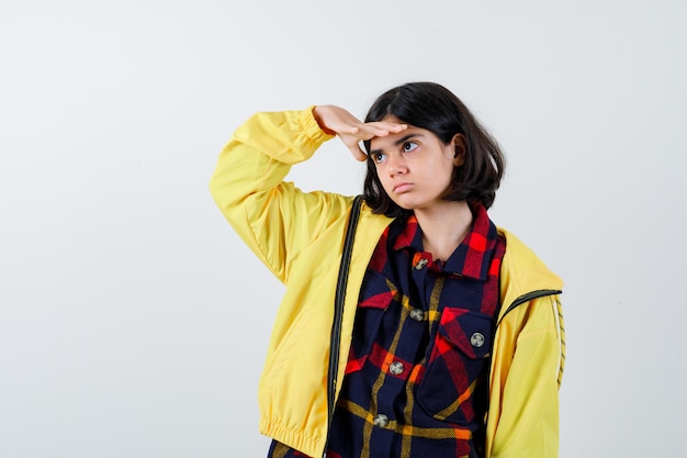 Niña con camisa a cuadros, chaqueta sosteniendo la mano en la frente en busca de manera y mirando enfocado, vista frontal.