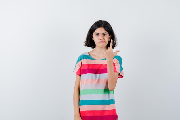 Niña en camiseta apuntando hacia arriba y mirando vacilante, vista frontal.