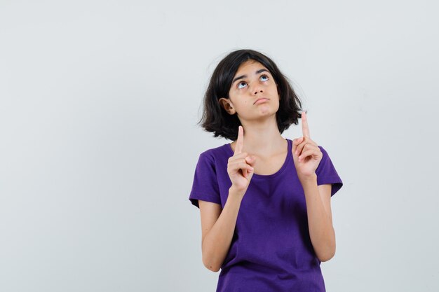 Niña en camiseta apuntando hacia arriba y mirando vacilante,