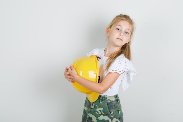 Niña en camiseta blanca, falda abrazando casco de seguridad y mirando confiada