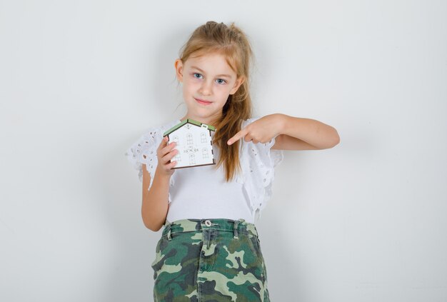 Niña en camiseta blanca, falda apuntando con el dedo al modelo de la casa y mirando esperanzado