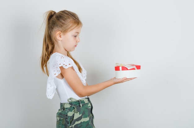 Niña en camiseta blanca, falda dando caja de regalo a alguien y mirando con cuidado