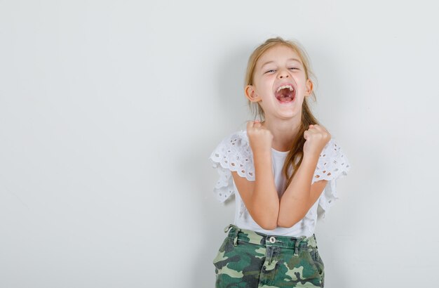 Niña en camiseta blanca, falda juntando las manos en puños para celebrar y lucir alegre