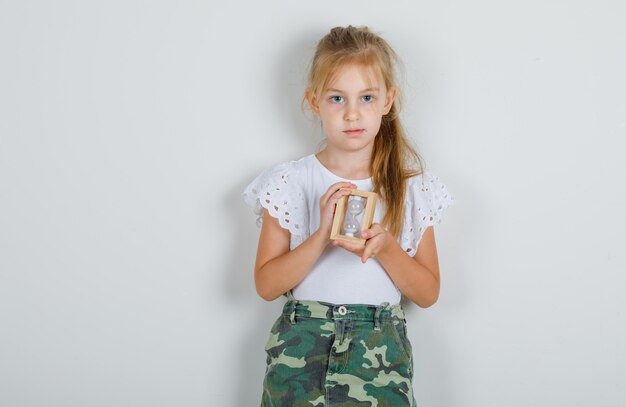 Niña en camiseta blanca, falda sosteniendo reloj de arena y mirando con cuidado