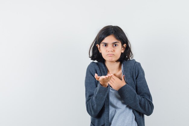 Niña en camiseta, chaqueta estirando la mano de manera interrogante y mirando perplejo, vista frontal.