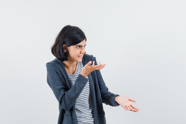 Niña en camiseta, chaqueta estirando las manos en gesto de interrogación y mirando curioso