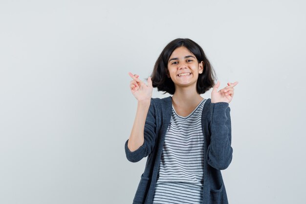 Niña en camiseta, chaqueta manteniendo los dedos cruzados y mirando contento,