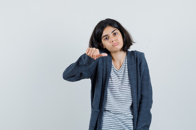 Niña en camiseta, chaqueta mostrando medio pulgar y mirando triste,