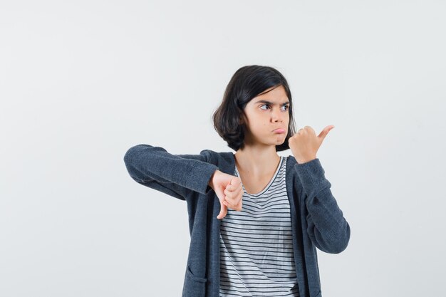 Niña en camiseta, chaqueta mostrando los pulgares hacia arriba y hacia abajo y mirando vacilante