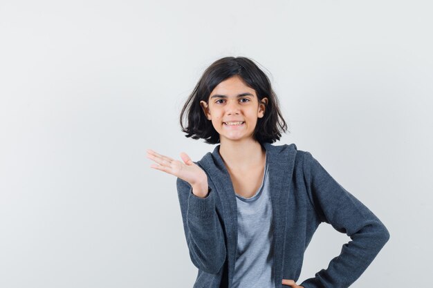 Niña en camiseta, chaqueta pretendiendo sostener o mostrar algo y mirando confiada, vista frontal.