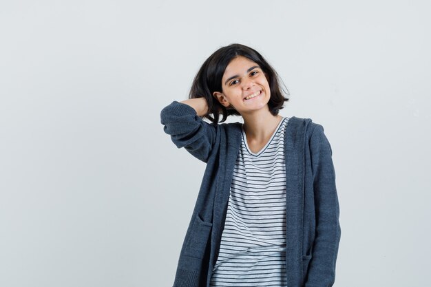 Niña en camiseta, chaqueta sosteniendo la mano en el cuello y mirando alegre,