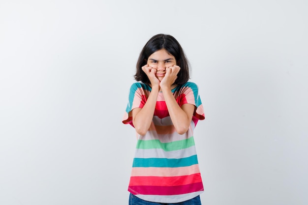 Niña en camiseta cogidos de la mano en las mejillas y mirando feliz, vista frontal.