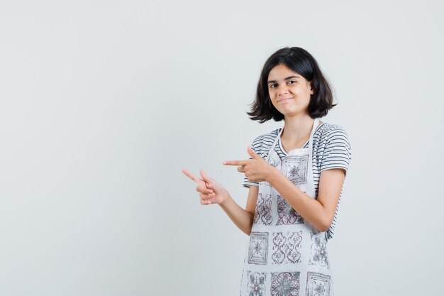 Niña en camiseta, delantal apuntando a un lado y mirando alegre,