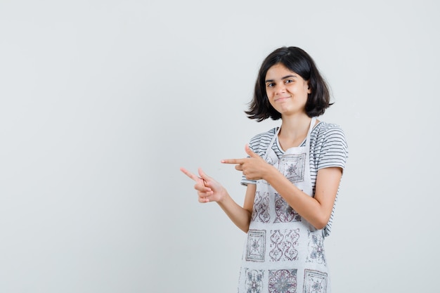 Niña en camiseta, delantal apuntando a un lado y mirando alegre,