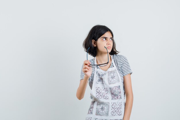 Niña en camiseta, delantal con gafas y mirando pensativo,