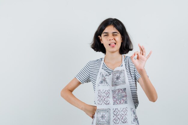 Niña en camiseta, delantal haciendo gesto bien, sacando la lengua, guiñando el ojo,