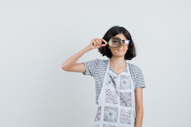 Niña en camiseta, delantal mirando a través de una lupa y luciendo sensato,