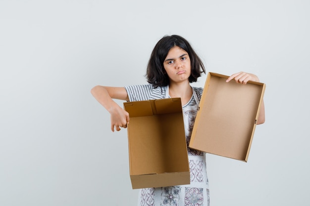 Niña en camiseta, delantal sosteniendo una caja de cartón vacía y mirando abatido,