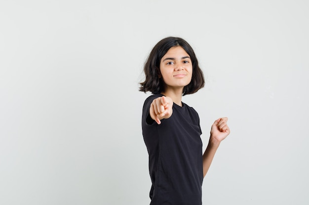 Foto gratuita niña en camiseta negra apuntando al frente y mirando alegre, vista frontal.