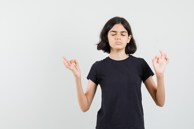 Niña en camiseta negra haciendo meditación con los ojos cerrados y mirando pacífica, vista frontal.