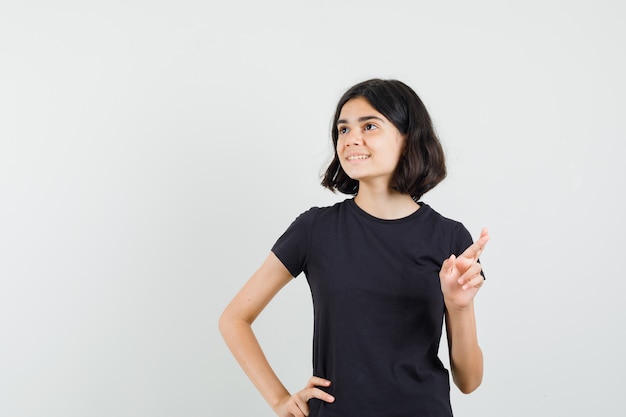 Niña en camiseta negra manteniendo los dedos cruzados y mirando esperanzado, vista frontal.