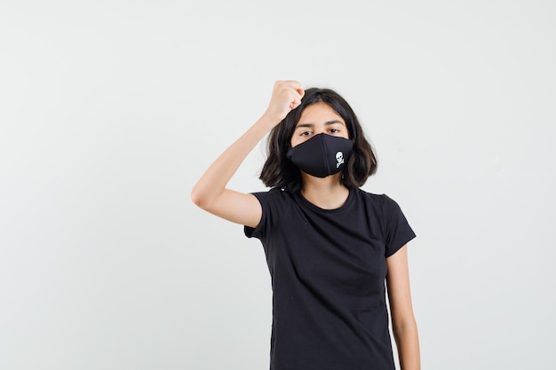 Foto gratuita niña en camiseta negra, máscara levantando el puño cerrado y mirando rencoroso, vista frontal.