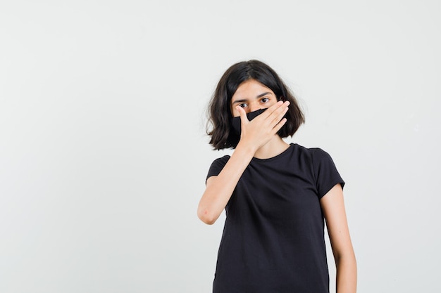 Foto gratuita niña en camiseta negra, máscara que cubre la boca con la mano, vista frontal.