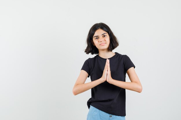 Niña en camiseta negra, pantalones cortos mostrando gesto de namaste y mirando esperanzado, vista frontal.