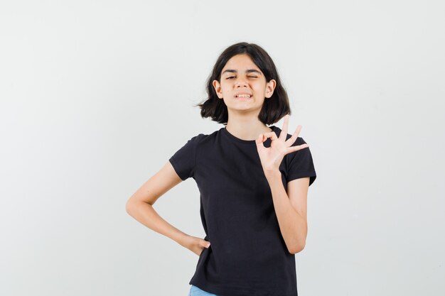 Niña en camiseta negra, pantalones cortos mostrando gesto ok, guiño de ojo, vista frontal.