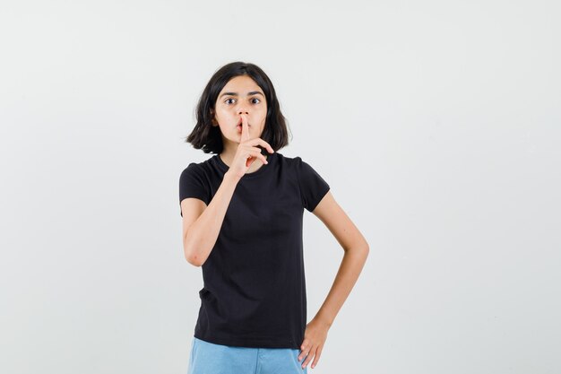 Niña en camiseta negra, pantalones cortos mostrando gesto de silencio y mirando con cuidado, vista frontal.