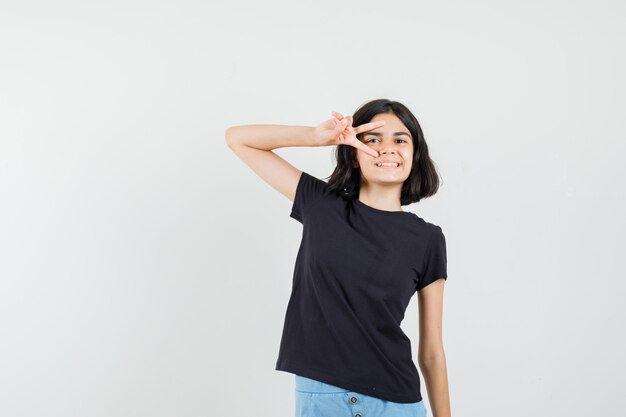 Niña en camiseta negra, pantalones cortos que muestran el signo v cerca del ojo y lucen alegre, vista frontal.