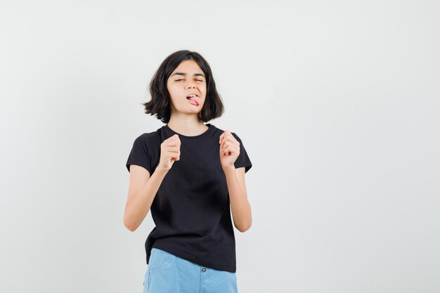 Niña en camiseta negra, pantalones cortos sacando la lengua y mirando divertido, vista frontal.