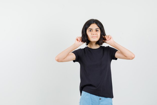 Niña en camiseta negra, pantalones cortos tapando las orejas con los dedos, vista frontal.