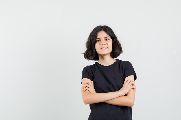 Niña en camiseta negra de pie con cruces de brazos y mirando confiado, vista frontal.