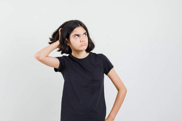 Niña en camiseta negra rascándose la cabeza y mirando pensativa, vista frontal.