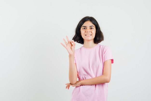 Niña en camiseta rosa haciendo bien gesto y mirando feliz, vista frontal.