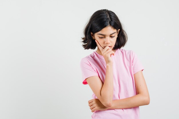 Niña en camiseta rosa mirando hacia abajo de pie en pose de pensamiento, vista frontal.