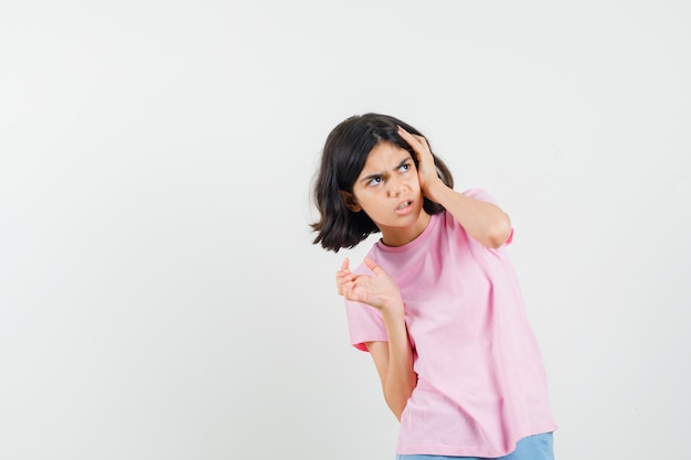 Niña en camiseta rosa, pantalones cortos sosteniendo la mano en la cabeza y mirando ofendido, vista frontal.