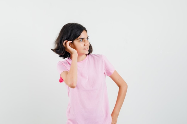 Niña en camiseta rosa sosteniendo la mano detrás de la oreja y mirando curiosa, vista frontal.