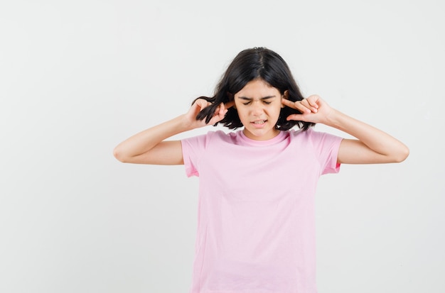 Niña en camiseta rosa tapando los oídos con los dedos y mirando molesto, vista frontal.