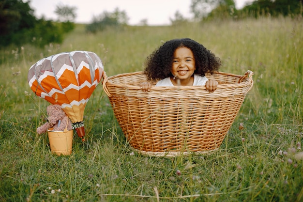 Niña en canasta de mimbre con globos a la luz del sol en verano. Niño feliz en el campo de la naturaleza sobre un césped