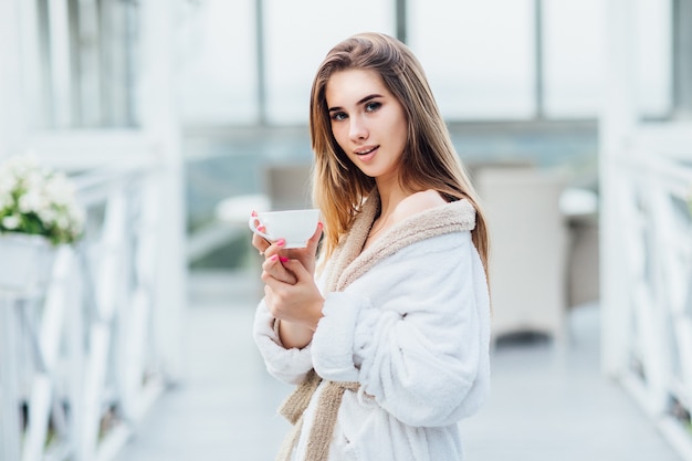 Una niña cansada de pie en la terraza con una hermosa vista y sosteniendo una taza.